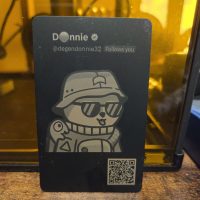 Black NFC card engraved with the Twitter profile of Donnie (@degendonnie32, verified). The card shows a cartoon character wearing a bucket hat and sunglasses. A QR code is printed at the bottom right.